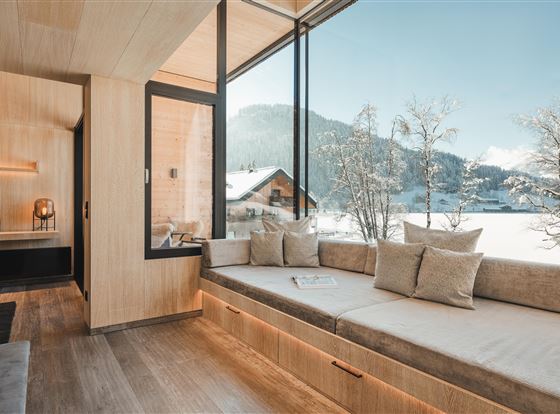 Ein modernes, helles Wohnzimmer mit großen Fenstern und einem Blick auf verschneite Berge. Der Sitzbereich ist gemütlich mit Kissen und holzverkleideten Wänden gestaltet.