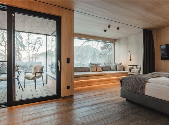 Ein modernes Hotelzimmer mit einem großen Fenster und Blick auf einen Balkon. Der Raum ist mit Holz und neutralen Farben eingerichtet und wirkt einladend.