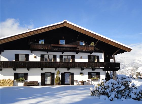 Ein schönes, alpenländisches Haus mit mehreren Balkonen, umgeben von Schnee. Der klare Himmel und die Berglandschaft verleihen der Szene eine ruhige Atmosphäre.