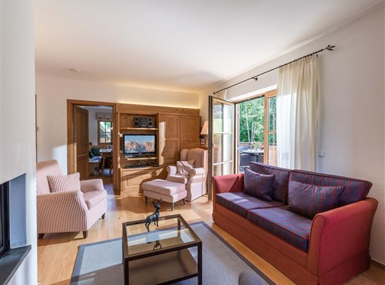 Ein gemütliches Wohnzimmer mit einem roten Sofa und zwei beigen Sessel. Große Fenster sorgen für viel Tageslicht und einen Blick ins Freie.