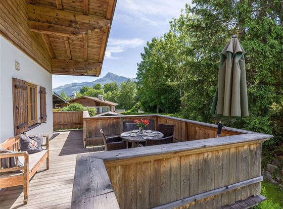 Eine gemütliche Terrasse mit Holzgeländer und einem Tisch. Im Hintergrund sind Bäume und Berge sichtbar.