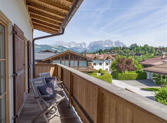 Ein schöner Balkon mit Holzmöbeln und Blick auf die Berge. Im Hintergrund sind grüne Wiesen und Häuser zu sehen.
