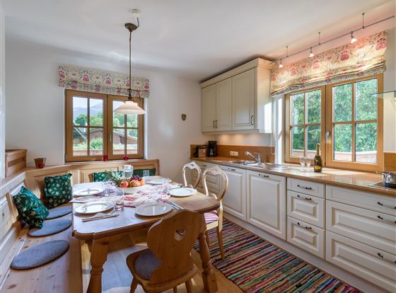 Eine einladende Küche mit einem Tisch, der für ein gemeinsames Essen gedeckt ist. Große Fenster lassen viel Licht herein und bieten einen Blick ins Grüne.