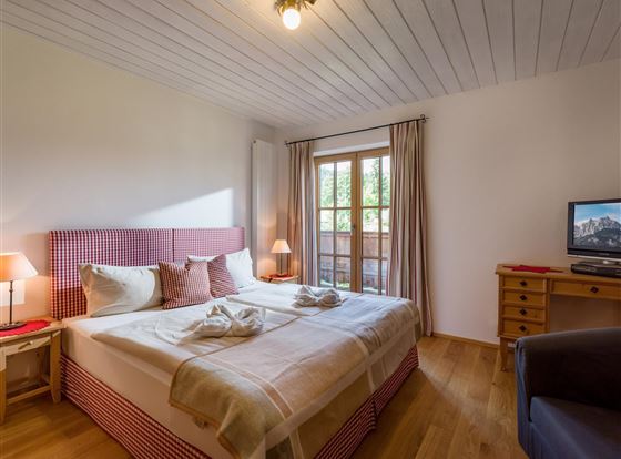 Ein gemütliches Schlafzimmer mit einem Doppelbett, Holzfußboden und großen Fenstern. Es gibt einen Fernseher und eine angenehme Atmosphäre.