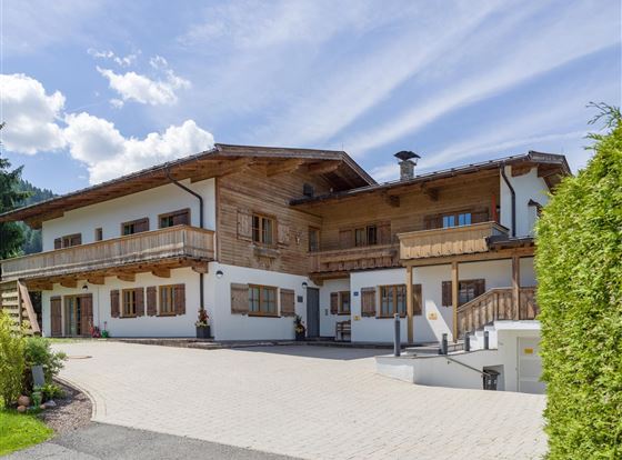 Ein modernes Haus im alpenländischen Stil mit Holzverkleidung. Der Außenbereich hat eine gepflasterte Einfahrt und eine grüne Hecke.