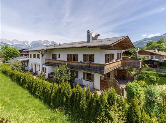 Ein schönes Haus im alpinen Stil mit Balkon und Garten. Im Hintergrund sind grüne Wiesen und Berge zu sehen.