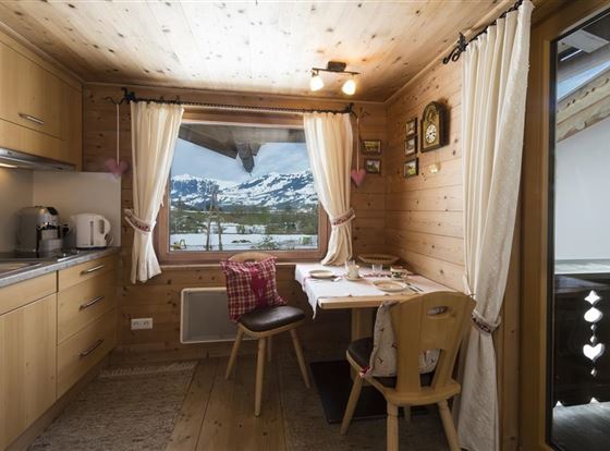 Eine gemütliche Küche mit Holzvertäfelung und einem Tisch, der auf eine schöne Aussicht auf die Berge hinausblickt. Der Raum hat helle Vorhänge und eine warme, einladende Atmosphäre.