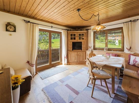 Ein gemütlicher Wohnraum mit Holzmöbeln und einem runden Esstisch. Große Fenster bieten einen Blick ins Grüne.