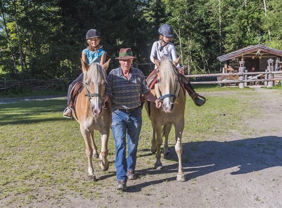 Ein Mann führt zwei Pferde, auf denen jeweils ein Kind sitzt. Im Hintergrund sind Bäume und ein Holzgebäude zu sehen.