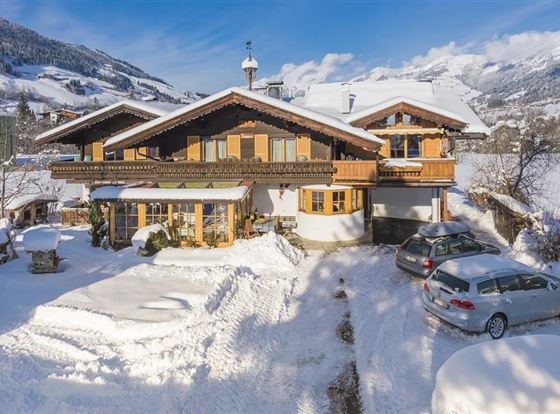 Ein großes, charmantes Holzhaus im Schnee mit braunen Fensterläden. Umgeben von schneebedeckten Bergen und einer ruhigen Winterlandschaft.