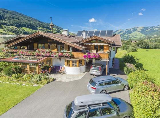Ein schönes Haus im alpinen Stil mit Blumenschmuck und Solarpanelen. Umgeben von saftigem Grün und Bergen im Hintergrund.