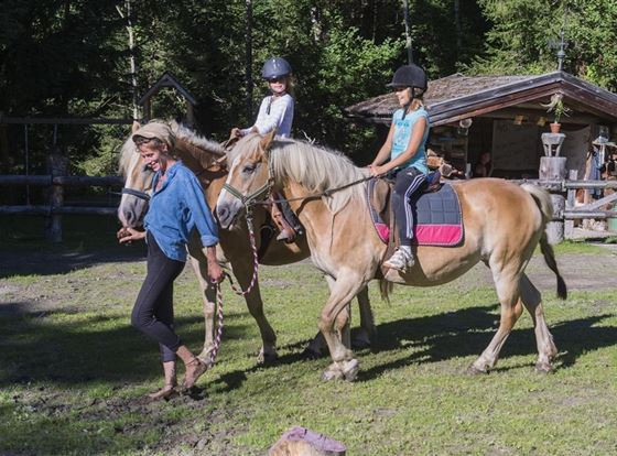 Zwei Mädchen reiten auf Pferden, während eine Frau sie führt. Im Hintergrund sind Bäume und eine Holzscheune sichtbar.