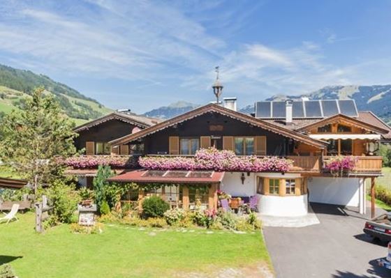 Ein charmantes alpenländisches Haus mit blühenden Pflanzen und einem schönen Garten. Im Hintergrund sind grüne Hügel und ein blauer Himmel zu sehen.