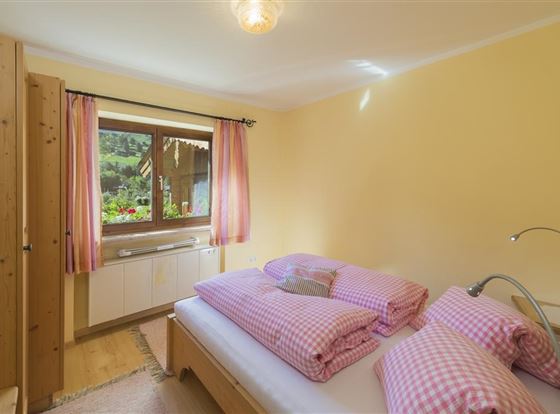 Ein gemütliches Schlafzimmer mit einem großen Fenster und Blick ins Grüne. Die Bettwäsche ist rosa kariert und die Wände sind in einem warmen Gelbton gestrichen.