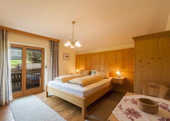 Ein helles Schlafzimmer mit einem großen Bett und Holzmöbeln. Der Raum hat Fenster mit Blick auf die Natur und einen gemütlichen Tisch.
