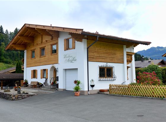 Ein modernes Haus im alpinen Stil mit Holzverkleidung und einem großen Balkon. Die schöne Umgebung wird von Bergen und grünen Feldern umgeben.