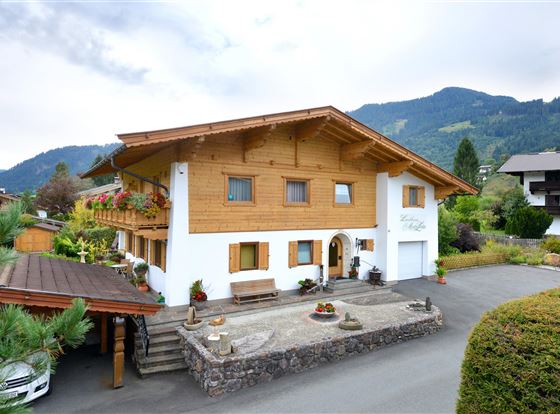 Ein charmantes, zweigeschossiges Haus mit Holzverkleidung und einem einladenden Eingang. Die umliegende Landschaft ist grün und bergig, was eine malerische Atmosphäre schafft.