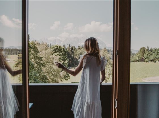 Eine Frau steht auf einem Balkon und blickt in die Natur. Im Hintergrund sind Bäume und Berge zu sehen.