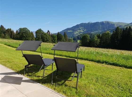 Zwei Liegestühle stehen auf einer Wiese mit Blick auf grüne Hügel und Bäume. Der Himmel ist klar und blau.