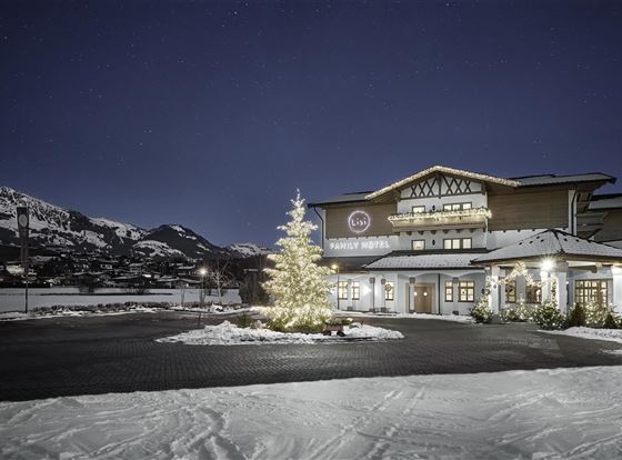 Ein Hotelgebäude im Schnee bei Nacht, umgeben von Bergen. Ein festlich geschmückter Weihnachtsbaum ist im Vordergrund zu sehen.