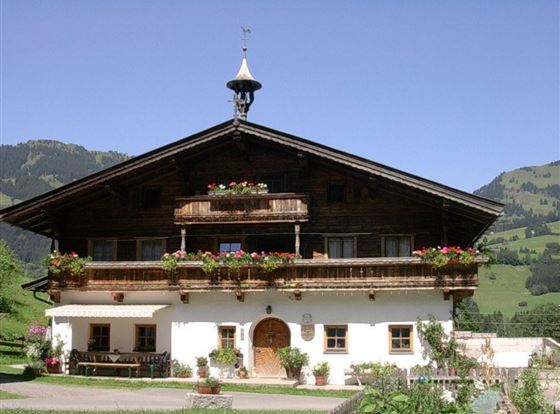 Ein traditionelles alpenländisches Haus mit Holzfassade und bunten Blumen entlang der Fenster. Im Hintergrund sind grüne Hügel und Berge zu sehen.