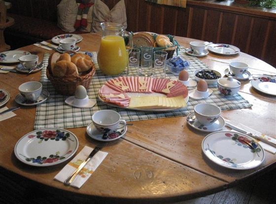 Ein rustikeller Tisch gedeckt für ein Frühstück. Es gibt Brötchen, Eier, Käse und einen Krug mit Saft.