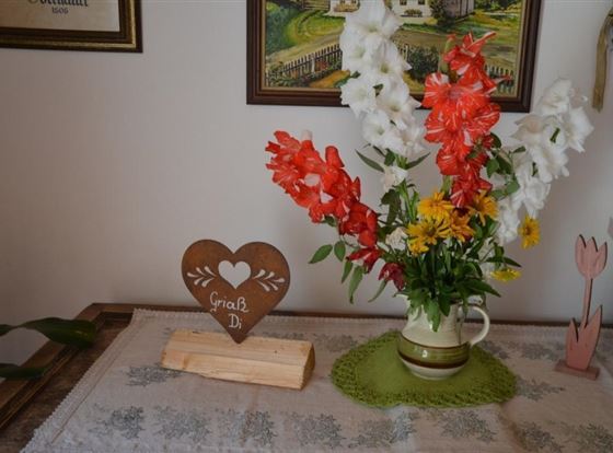 Ein hübscher Blumenstrauß aus roten, weißen und gelben Blumen in einer Kanne steht auf einem Tisch. Daneben liegt ein herzförmiges Holzschild mit der Aufschrift "Gruß Di".