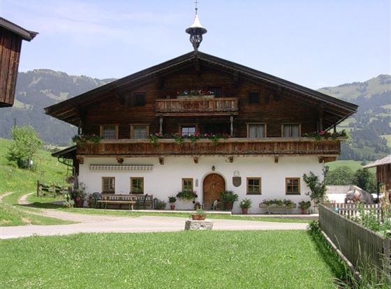 Ein traditionelles alpenländisches Haus mit Holzverkleidung und hübschen Balkonen. Umgeben von grünen Wiesen und Bergen im Hintergrund.