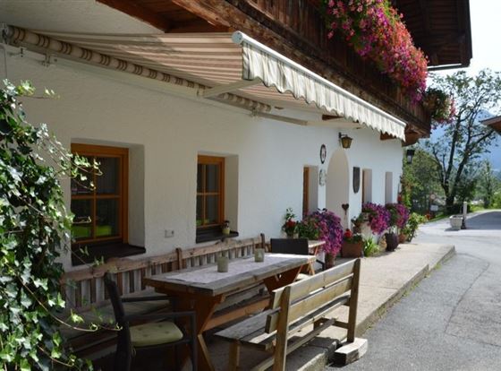Eine charmante Almhütte mit bunten Blumen und einer gemütlichen Sitzecke. Der Weg führt durch eine malerische Umgebung mit sanften Hügeln.