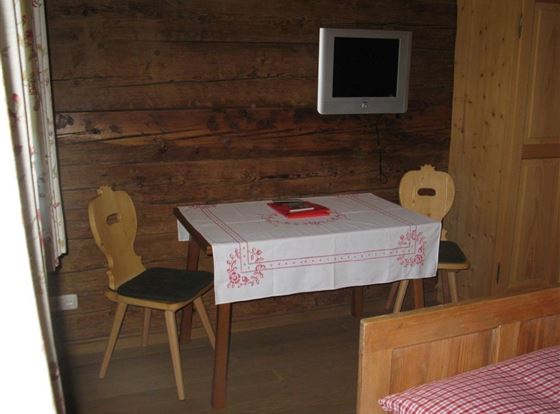 Ein einfach eingerichteter Raum mit einem Holztisch und zwei Stühlen. An der Wand hängt ein Fernseher, und der Boden ist aus Holz.