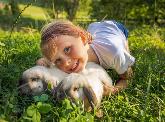 Ein fröhliches Kind liegt auf dem Gras und kuschelt mit zwei weißen Kaninchen. Die Sonne scheint und die Szene wirkt freundlich und entspannt.