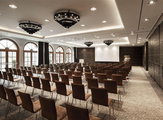 An elegant conference room with rows of chairs and a large screen. Large windows provide plenty of natural light.