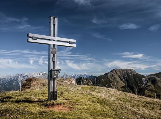 Ein großes, modernes Kreuz steht auf einem grünen Gipfel. Im Hintergrund erstrecken sich majestätische Berge unter einem klaren Himmel.