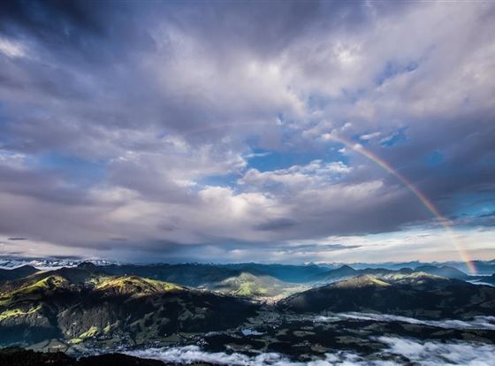Eine beeindruckende Berglandschaft unter einem bewölkten Himmel mit einem Regenbogen. Die Wolken und die Sonne schaffen ein malerisches Lichtspiel über den Tälern.
