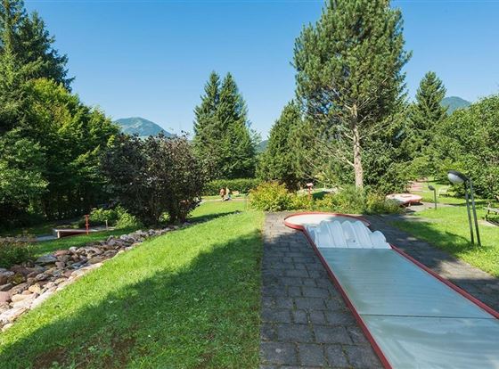 Eine grüne Gartenlandschaft mit Bäumen und Büschen. In der Mitte verläuft eine Rutsche auf einem gepflasterten Weg.