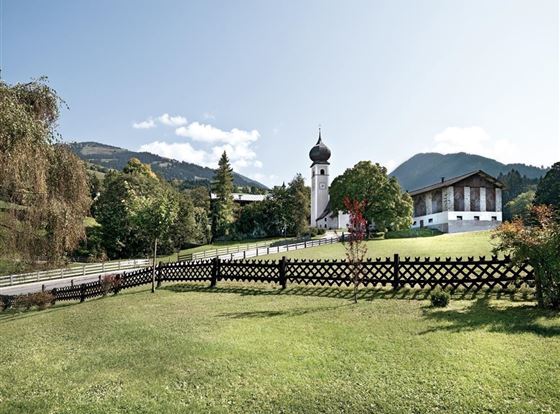 Eine ruhige Landschaft mit einem gepflegten Garten und einer traditionellen Kirche im Hintergrund. Die Umgebung ist von Hügeln und Bäumen umgeben, und der Himmel ist teils bewölkt.