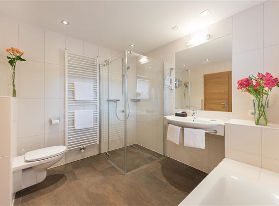 Ein modernes Badezimmer mit einer Dusche und einem Badewanne. Helle Fliesen und frische Blumen sorgen für eine einladende Atmosphäre.