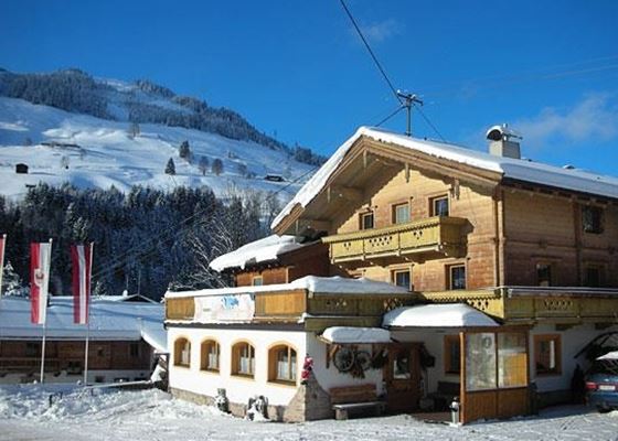 Ein gemütliches Holzhaus im Schnee mit blauen Himmel im Hintergrund. Die Umgebung ist von schneebedeckten Bergen und Tannenbäumen geprägt.