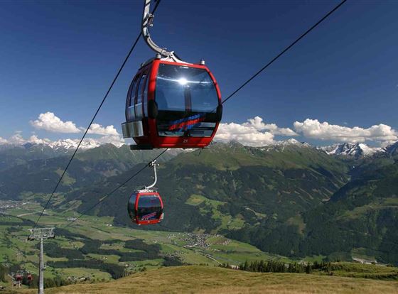 Eine Seilbahn fährt über eine grüne Berglandschaft. Im Hintergrund sind schneebedeckte Berge und ein klarer blauer Himmel zu sehen.