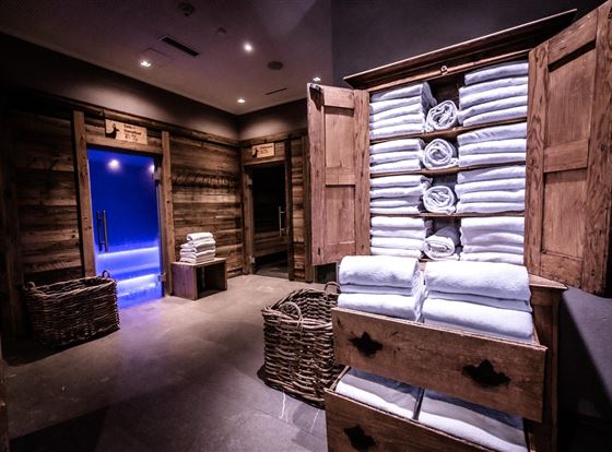 Ein stilvolles Spa mit einem Holzschrank voller gefalteter Handtücher. Im Hintergrund sind zwei Türen sichtbar, die zu einem Wellnessbereich führen.