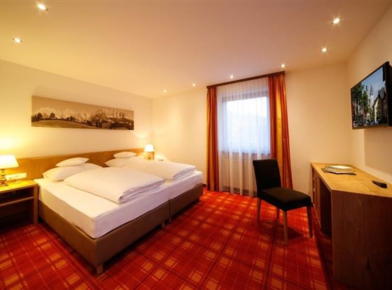 Ein modernes Hotelzimmer mit einem Doppelbett, einer Sitzgelegenheit und einem Fernseher. Das Zimmer verfügt über große Fenster mit Vorhängen und eine warme, einladende Atmosphäre.