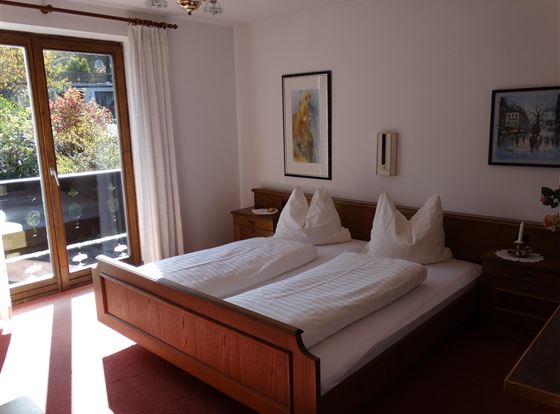 Ein gemütliches Schlafzimmer mit einem Doppelbett und holzene Möbel. Große Fenster lassen viel Tageslicht hereinfällt und bieten einen Blick auf den Balkon.