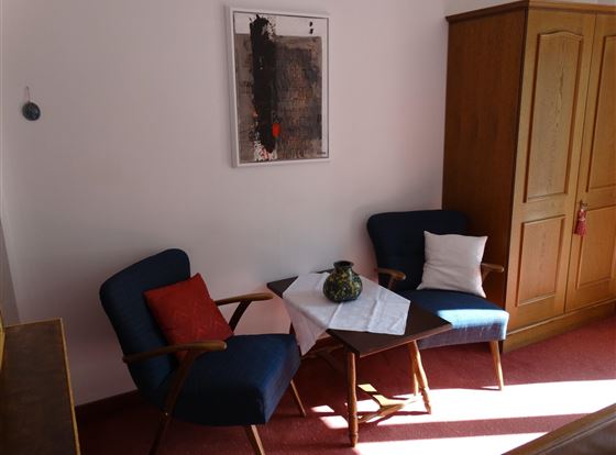 Ein gemütlicher Wohnbereich mit zwei blauen Stühlen und einem kleinen Tisch. An der Wand hängt ein modernes Kunstwerk, und ein Schrank steht rechts im Bild.