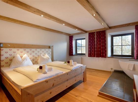 Ein gemütliches Schlafzimmer mit einem Doppelbett aus Holz und weißen Kissen. Große Fenster lassen viel Licht hereinfliessen und schaffen eine einladende Atmosphäre.