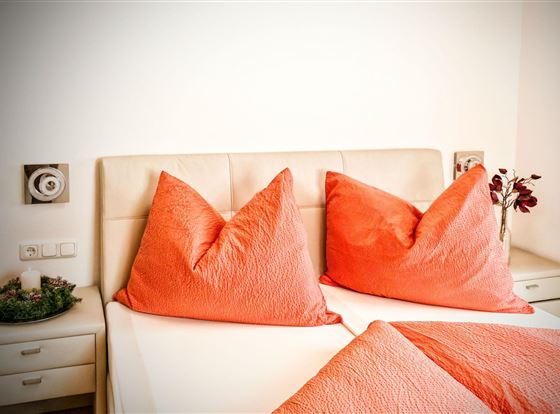 Ein gemütliches Schlafzimmer mit einem großen Bett und auffälligen, orangen Kissen. An der Wand sind dekorative Elemente und ein kleines Arrangement von Blumen zu sehen.