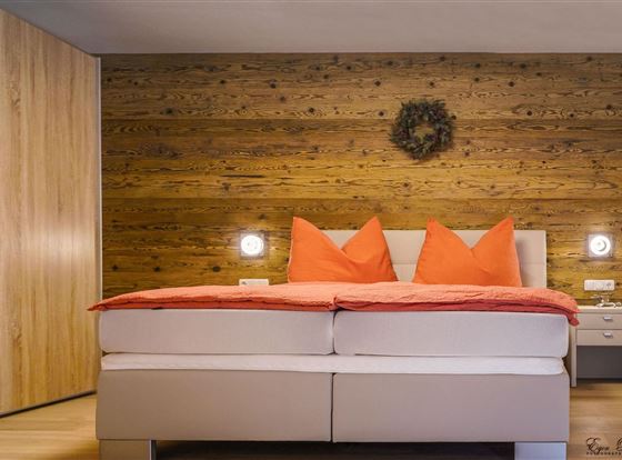 Ein modernes Schlafzimmer mit einem großen Bett und leuchtend orangefarbenen Kissen. Die Wände sind aus Holz, und es gibt eine stilvolle Nachtkommode.