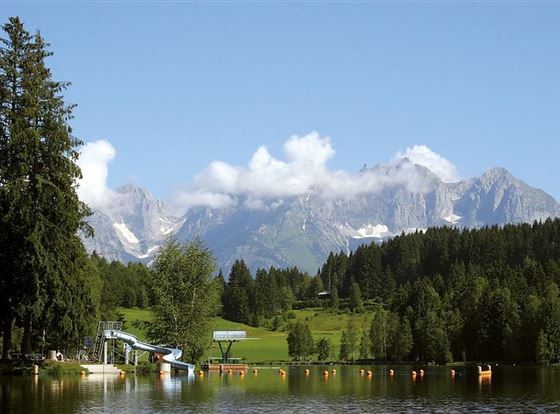 Ein ruhiger See umgeben von grünen Bäumen und beeindruckenden Bergen im Hintergrund. Der Himmel ist klar und blau mit einigen Wolken.
