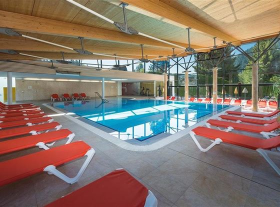 Ein modernes Schwimmbad mit einem glasklaren Wasser. Rote Liegen säumen den Poolbereich und große Fenster sorgen für viel Licht.