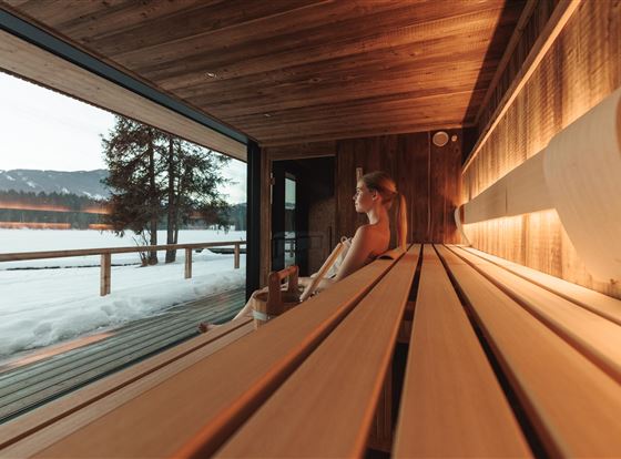 Ein modernes Sauna-Innenraum mit Holzregalen und großen Fenstern. Im Hintergrund ist eine winterliche Landschaft mit Schnee und Bäumen zu sehen.
