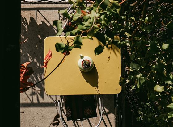 Ein kleiner Tisch mit einer bunten Schale steht im Freien, umgeben von Pflanzen. Das Sonnenlicht fällt auf die Tischplatte und sorgt für eine einladende Atmosphäre.
